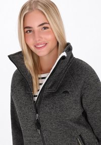 Siva zip-up jakna z visokim ovratnikom, izdelana iz teksturirane tkanine, z black dodatki in fleecem obloženim notranjim delom. Pokaže se prugasta majica pod njo.