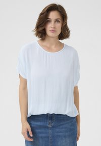 Blouse bleu clair, ample, à manches courtes, avec un col rond et une texture froissée, associée à une jupe en denim.