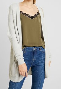 Světle šedý kardigan s vroubkovanou texturou, nošený přes olivově zelený top s černou krajkou. V kombinaci s modrými denimovými džíny.