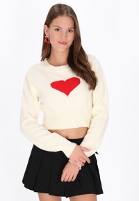 Crème cropped trui met een rood hartontwerp. Heeft lange mouwen en geribbelde manchetten. Gepaard met een zwarte plooienrok.