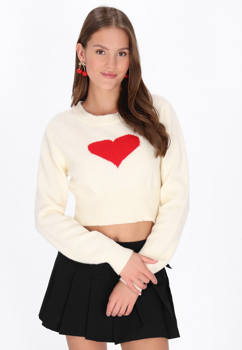 Crème cropped trui met een rood hartontwerp. Heeft lange mouwen en geribbelde manchetten. Gepaard met een zwarte plooienrok.