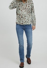 Blouse à manches longues vert clair avec un col en V, présentant un motif floral marron et noir. Associée à un jean bleu et des bottines marron.