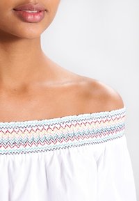 Primo piano della spalla e delle labbra di una persona che indossa una blusa bianca con spalle scoperte, decorata con ricami colorati a zigzag.
