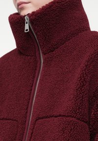 Prim-plan al unei persoane îmbrăcate cu o geacă din fleece de culoare vișinie, cu fermoar, guler înalt și țesătură texturată, pufoasă.