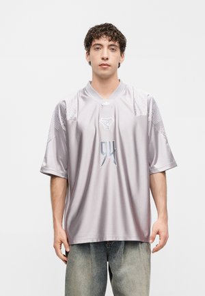 Nuori mies, jolla on kiharat hiukset, päällään löysä hopeanharmaa urheilupaita numerolla 94 ja vaaleaksi pestyt farkut, seisoo yksiväristä taustaa vasten.