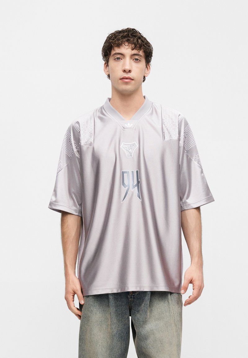 Νέος άνδρας με σγουρά μαλλιά που φοράει χαλαρό ασημί-γκρι αθλητικό μπλουζάκι με τον αριθμό 94 και ελαφρώς ξεβαμμένα τζιν, στέκεται μπροστά σε απλό φόντο.