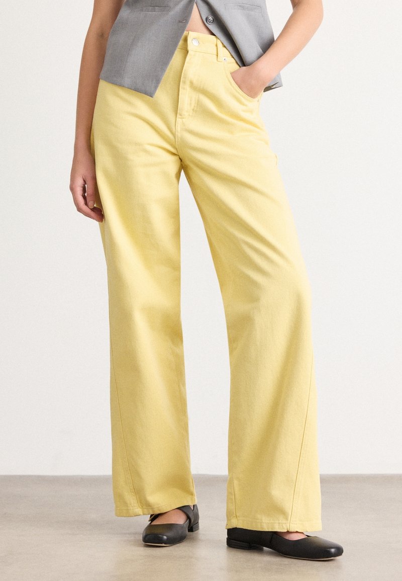 Personne portant un pantalon jaune taille haute à jambes larges, des chaussures plates noires et un blazer gris court, main dans la poche, debout sur un sol gris.
