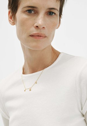 Primer plano de una persona con cabello corto y color castaño que lleva una camisa blanca y un delicado collar de oro con pequeños colgantes en forma de estrella.