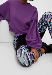 Sudadera morada, recortada, con mangas globosas combinada con leggins multicolores y estampados que presentan tonos verdes, morados y acentos en negro.