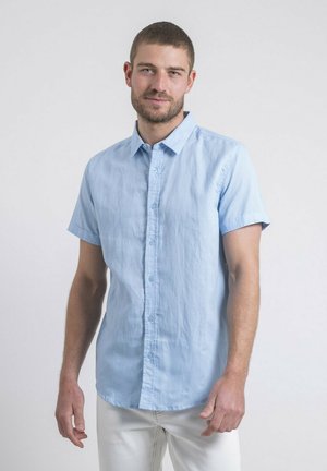 Homme aux cheveux bruns courts et barbe, portant une chemise bleu clair à manches courtes boutonnée et un pantalon blanc, debout devant un fond uni.