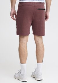 Shorts bordeaux en tissu doux avec un design ajusté, dotés d'une poche zippée au dos et d'ourlets. Portés avec des chaussettes blanches et des baskets.