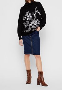 Zwarte oversized trui met zilveren bloemdecoratie, gecombineerd met een denim kokerrok en bruine enkellaarsjes met hak en glanzende textuur.