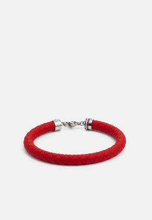 Červený pletený kožený náramek se stříbrnou sponou a kovovými zakončeními, zobrazený na jednobarevném bílém pozadí.