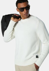 Maglione bianco a maglia con dettagli a coste sulle maniche e in vita, caratterizzato da una patch con logo nera in basso a sinistra. Indossato con una borsa nera.