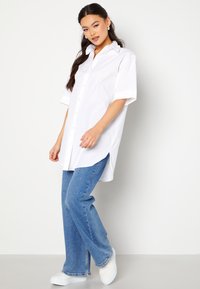 Camicia bianca a maniche corte con colletto e spacchi laterali, indossata su jeans a zampa blu e sneakers bianche. Tessuto morbido, vestibilità rilassata.