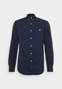 Chemise bleu marine à manches longues avec boutons sur le devant, col boutonné et petit logo brodé jaune sur le côté gauche de la poitrine.