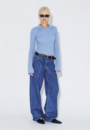 Person med kort blondt hår iført sorte solbriller, ribbet lyseblå knapbluse, vidde blå jeans, sort bælte og sorte spidse sko.