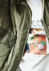 Weißes Hemd mit floralem Print und dem Text "MIT ROSEN UND ANEMONEN 1890", teilweise von einer offenen olivgrünen Steppjacke verdeckt.