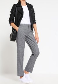 Kvinna som bär en svart läderjacka, vit t-shirt, grå rutiga byxor, vita sneakers och bär en svart axelväska, står med en hand i fickan.