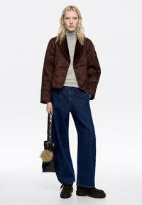 Młoda kobieta z długimi blond włosami, ubrana w brązową kurtkę, szary golf, szerokie niebieskie jeansy, ciemne buty, trzymająca czarną torbę na ramię z futrzanym pomponem.