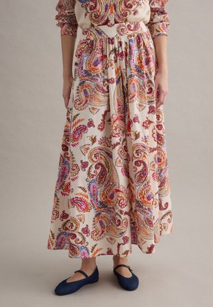 Donna che indossa una lunga gonna beige con fantasie paisley colorate rosa, arancione e viola, abbinata a ballerine Mary Jane blu navy.