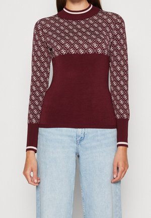 Femme portant un pull bordeaux avec des motifs géométriques blancs sur la partie supérieure de la poitrine et les manches, associé à un jean bleu clair.