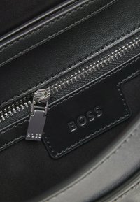 Mustat vetoketjut nahkalaukun sisällä, jossa on suorakaiteen muotoinen, kohokuvioitu "BOSS" -merkki. Sisältää sileän pinnan ja kiiltävää hopeista metallia.