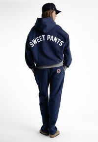 Ensemble de sweat à capuche bleu marine et de pantalon de jogging avec le texte "SWEET PANTS" en blanc, des accents gris et un patch logo circulaire sur le pantalon.