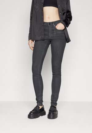 Frau trägt schwarzes kurzes Top, offene schwarze Bluse, dunkelgraue Skinny-Jeans und klobige schwarze Loafers, steht mit einer Hand in der Tasche.