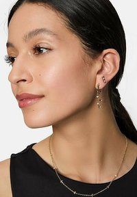 Orecchini a cerchio in oro con perline pendenti, abbinati a una delicata collana d'oro con piccoli anelli e dettagli, indossati da una donna.