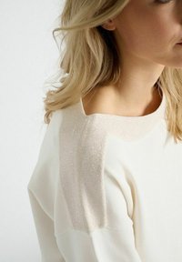 Crème-kleurige trui met een zachte, gestructureerde glinsterende beige schouderaccent. Heeft een ronde halslijn en een relaxte pasvorm.