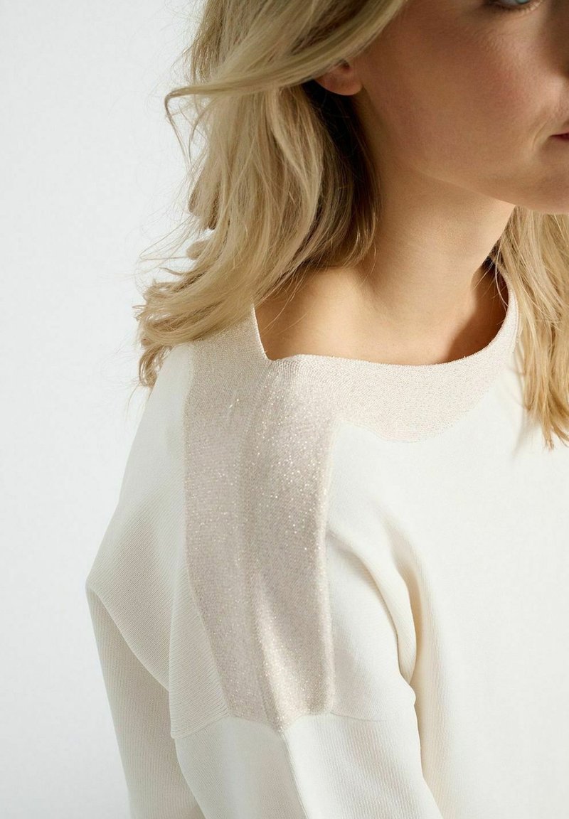 Crème-kleurige trui met een zachte, gestructureerde glinsterende beige schouderaccent. Heeft een ronde halslijn en een relaxte pasvorm.