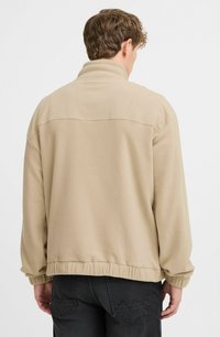 Beige fleecejakke med høj krave og elastiske manchetter. Har en ribbet nederdel og en glat, blød tekstur. Ingen synlig branding eller mønstre.