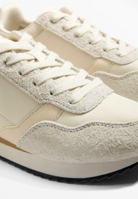 Beige träningsskor med texturerat mocka och slät läderöverdel, rund tå och cremefärgade snören; subtila vågiga detaljer längs sulkanten.