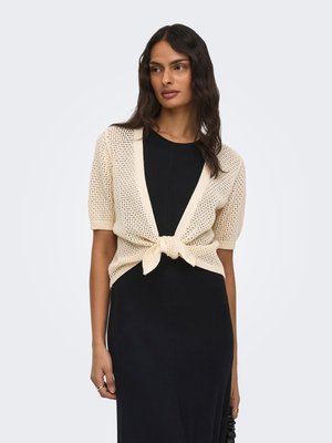 Femeie cu păr lung și închis la culoare, purtând o rochie neagră și un cardigan scurt bej tricotat, legat în față, pe un fundal simplu și deschis la culoare.