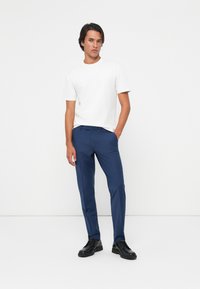 Junger Mann steht mit einer Hand in der Tasche, trägt ein weißes T-Shirt, blaue Hosen und schwarze Businessschuhe vor einem schlichten weißen Hintergrund.