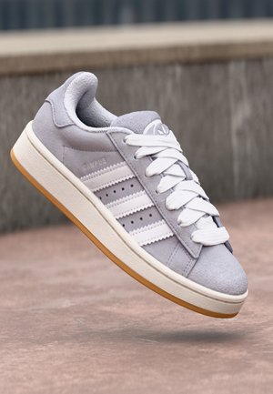 Jasnoszare buty sportowe Adidas Campus z białymi paskami, grubą białą podeszwą i gumową powierzchnią zewnętrzną, przedstawione w powietrzu nad betonową powierzchnią.