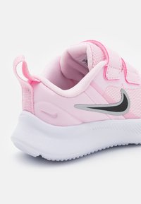 Zapatilla deportiva rosa con parte superior texturizada, dos correas de Velcro, un logo de Nike gris y una suela blanca. Forma redondeada con una lengüeta en la parte trasera.