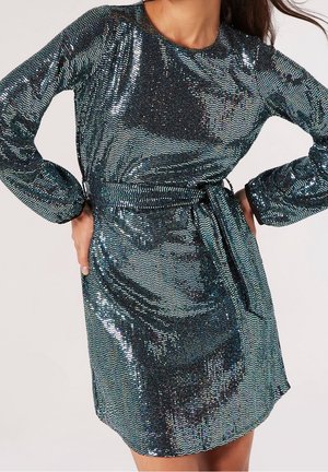 Kobieta w długim rękawie, sukience do kolan w kolorze metalicznej ciemnej zieleni z cekinami i paskiem związanym w talii, stojąca z rękami na biodrach.