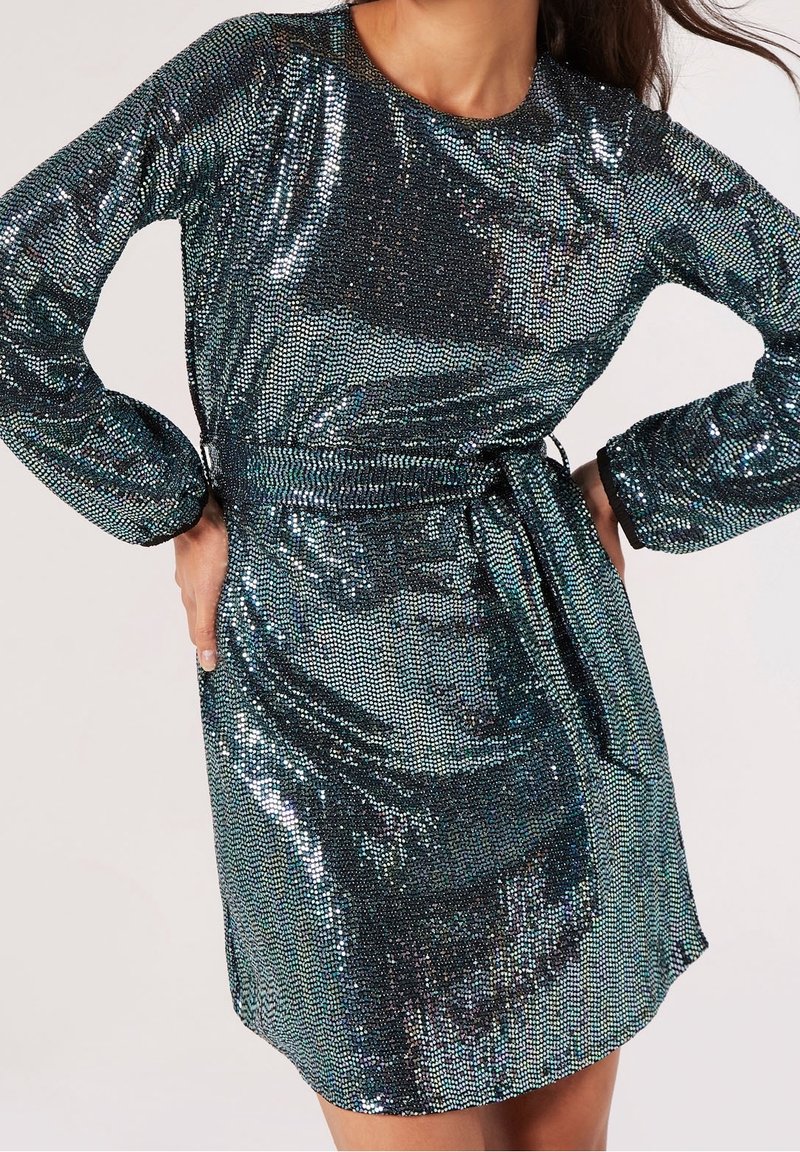 Femme portant une robe à manches longues, longueur genou, en sequins métalliques vert foncé avec une ceinture nouée à la taille, debout les mains sur les hanches.