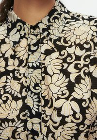 Camicia nera con bottoni, caratterizzata da un motivo floreale in bianco e giallo. Il tessuto ha un aspetto texturizzato e leggermente trasparente.
