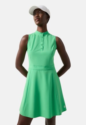 Woman wearing sleeveless bright green dress with buttoned collar and white cap, standing with hands behind her back.