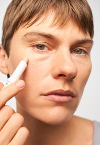 A white, twist-up concealer stick is applied to the under-eye area. Features smooth, skin-like texture, subtle beige color, and minimalistic design.