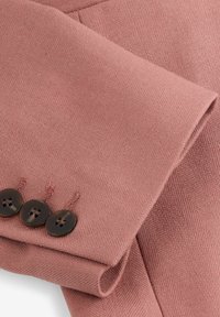 Růžový látkový materiál s texturovaným povrchem; má přehnutý manžet a tři tmavé knoflíky na okraji.