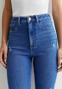 Jeans de denim azul de cintura alta con un acabado desgastado, pequeños desgarros y costuras en contraste. Diseño clásico de cinco bolsillos con cierre de botón metálico.