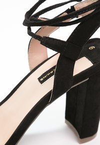 Czarne zamszowe sandały na wysokim obcasie z krzyżującymi się paskami, masywnym obcasem i gładką, nagą wkładką z złotym brandingiem.