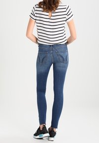 Vaqueros ajustados azules con bolsillos traseros y una camiseta de rayas de manga corta. Zapatillas negras con detalles azules. Vista trasera contra un fondo liso.