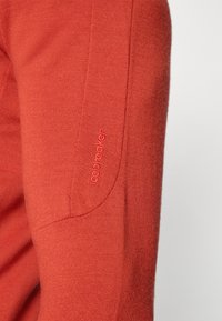 Verbrande oranje trui met een geribbelde textuur, voorzien van een afgeronde schoudernaad en een subtiele geborduurde logo op de mouw.