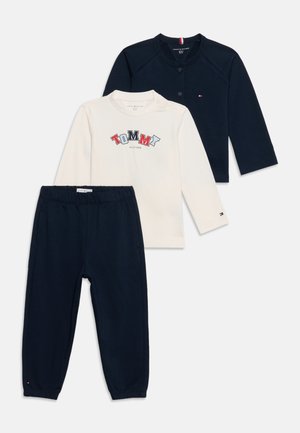 Granatowe spodnie z elastycznym pasem, kremowa koszula z długim rękawem z logo "Tommy" oraz granatowy kardigan zapinany na guziki z małą naszywką w kształcie flagi.