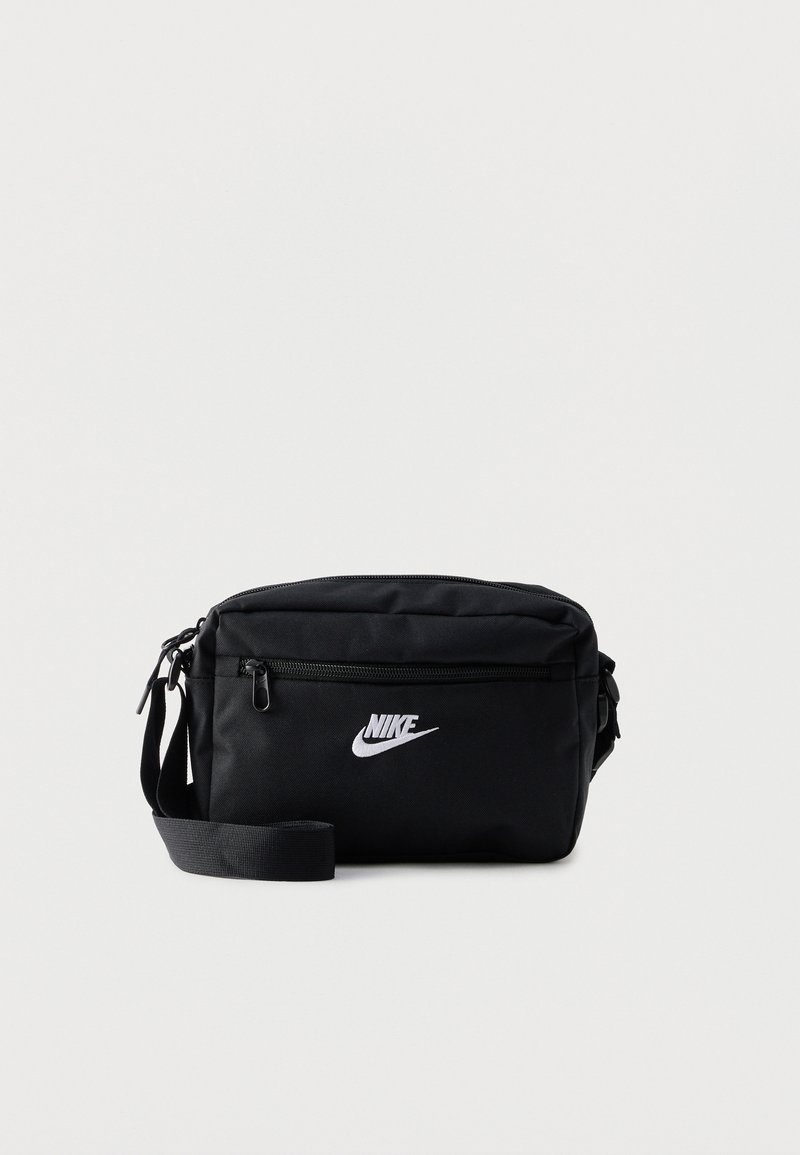 Schwarze Nike Crossbody-Tasche mit vorderer Reißverschlusstasche, verstellbarem Riemen und weißem Logo vorne vor einfarbigem Hintergrund.